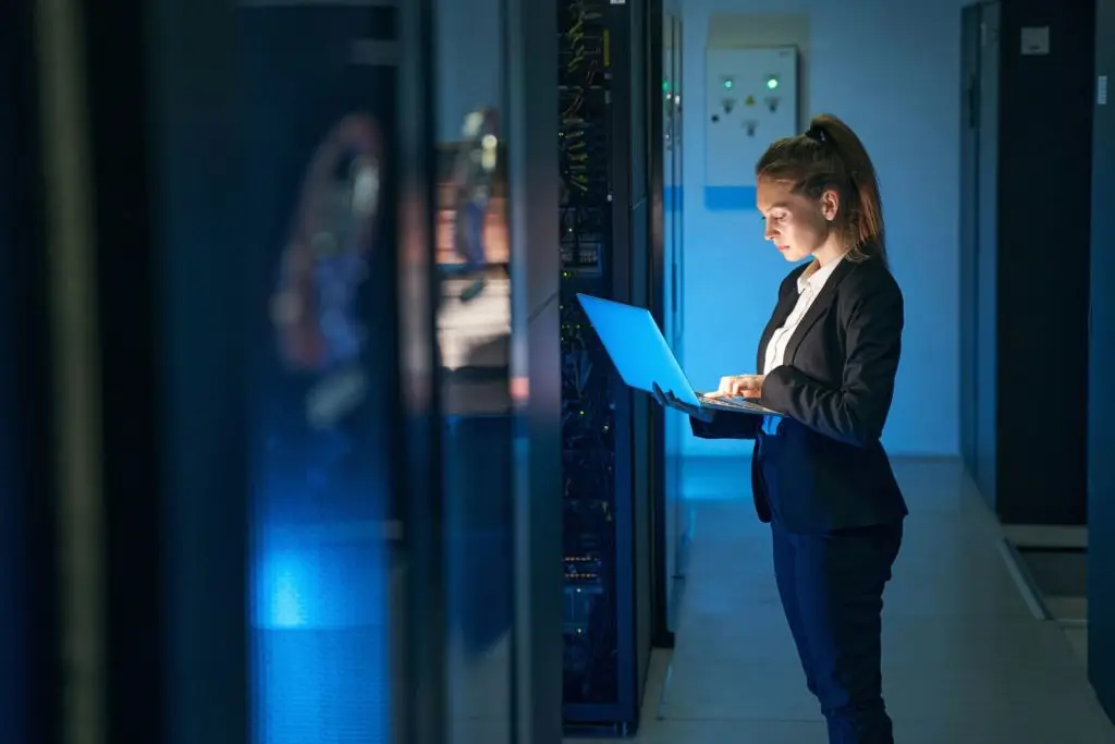A woman in a server room working on a laptop, surrounded by racks of computer servers. She is dressed in business attire and is illuminated by the blue light from the equipment.