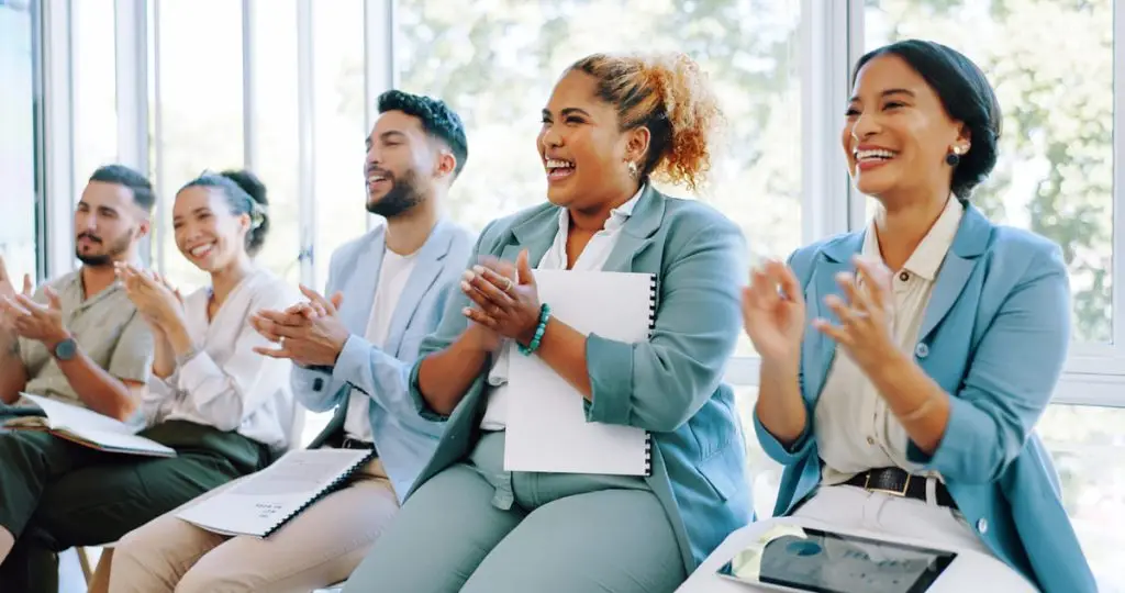A diverse group of business professionals joyfully clapping during a seminar. The focus is on an African American woman in the center, smiling broadly and applauding, dressed in a light blue blazer and holding a white notebook. She is surrounded by colleagues who are also engaged and clapping, with large windows allowing natural light to fill the room.