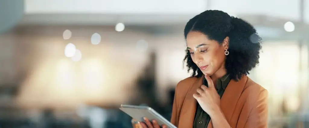A woman in a business suit, thoughtfully looking at a tablet in a well-lit, modern office space. The background is blurred, highlighting her focus on the device.